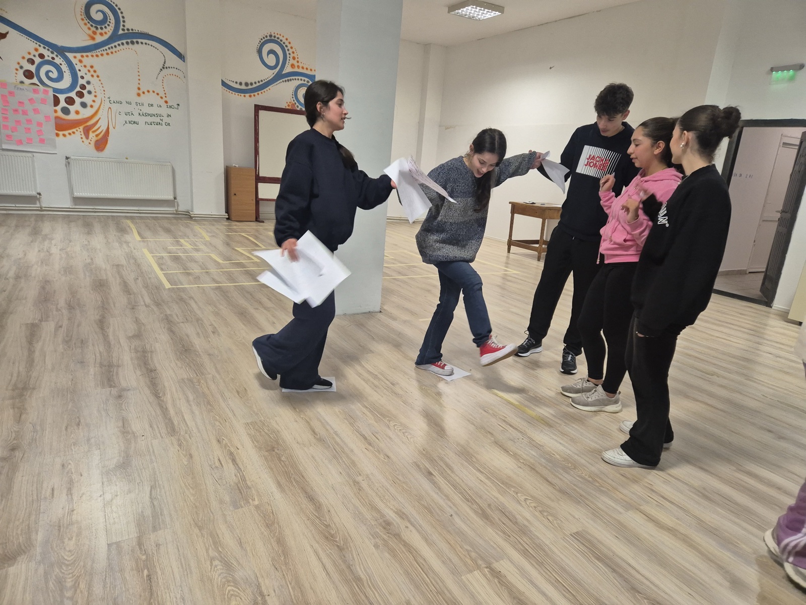A participant balances mid-step across paper stones while her team cheers